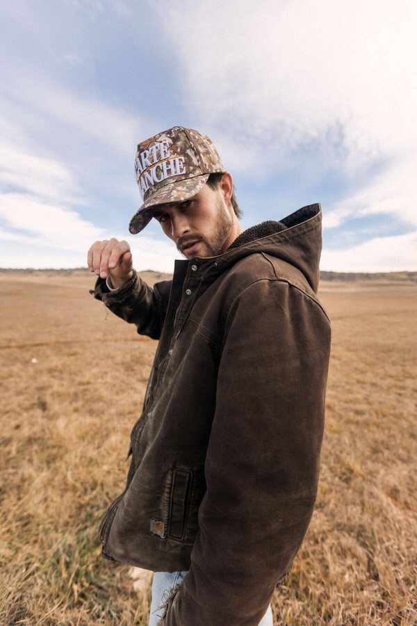Model in a camo Carte Blanche cap in an open field, 012 campaign frame